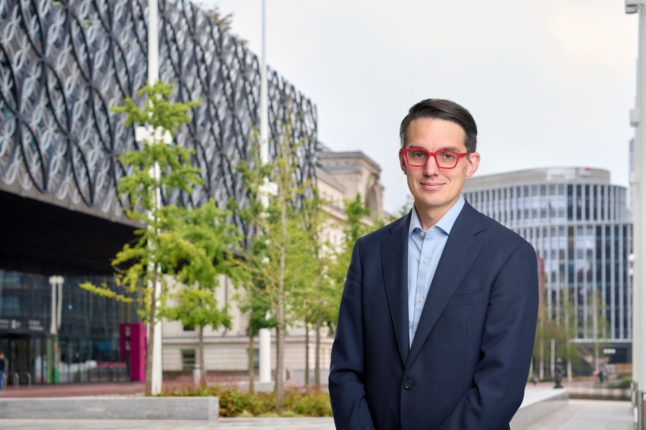 A man in a light blue shirt and suit jacket in front of some trees and buildings in central Birmingham.