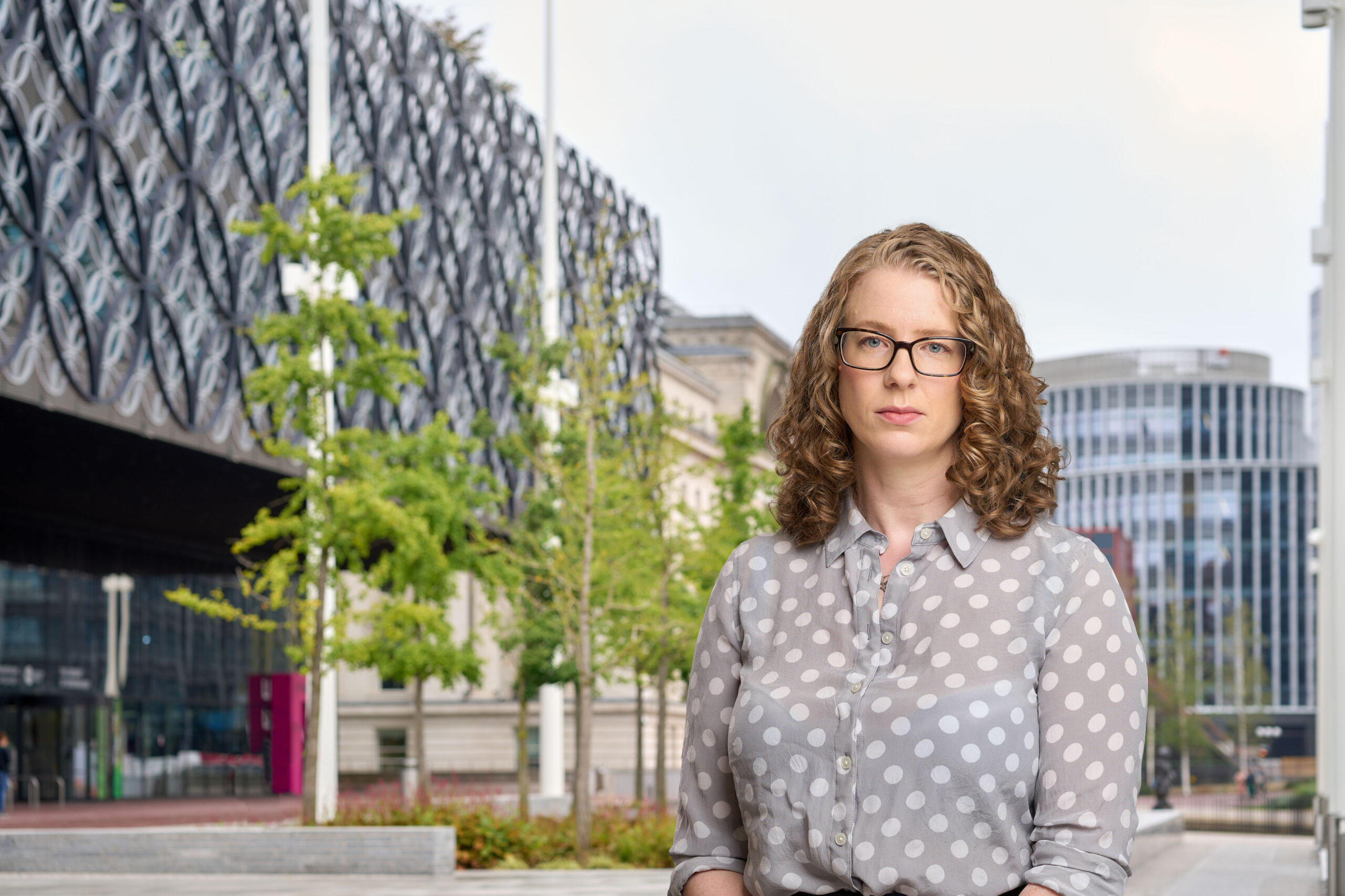 A woman in a light grey shirt with white polka dots in front of some trees and buildings in central Birmingham.