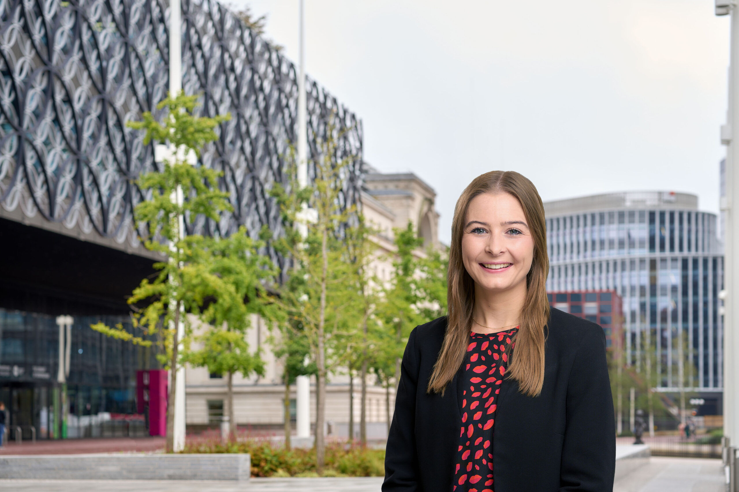 A woman in a red and black blouse and black blazer is standing and smiling in front of some trees and buildings in central Birmingham.