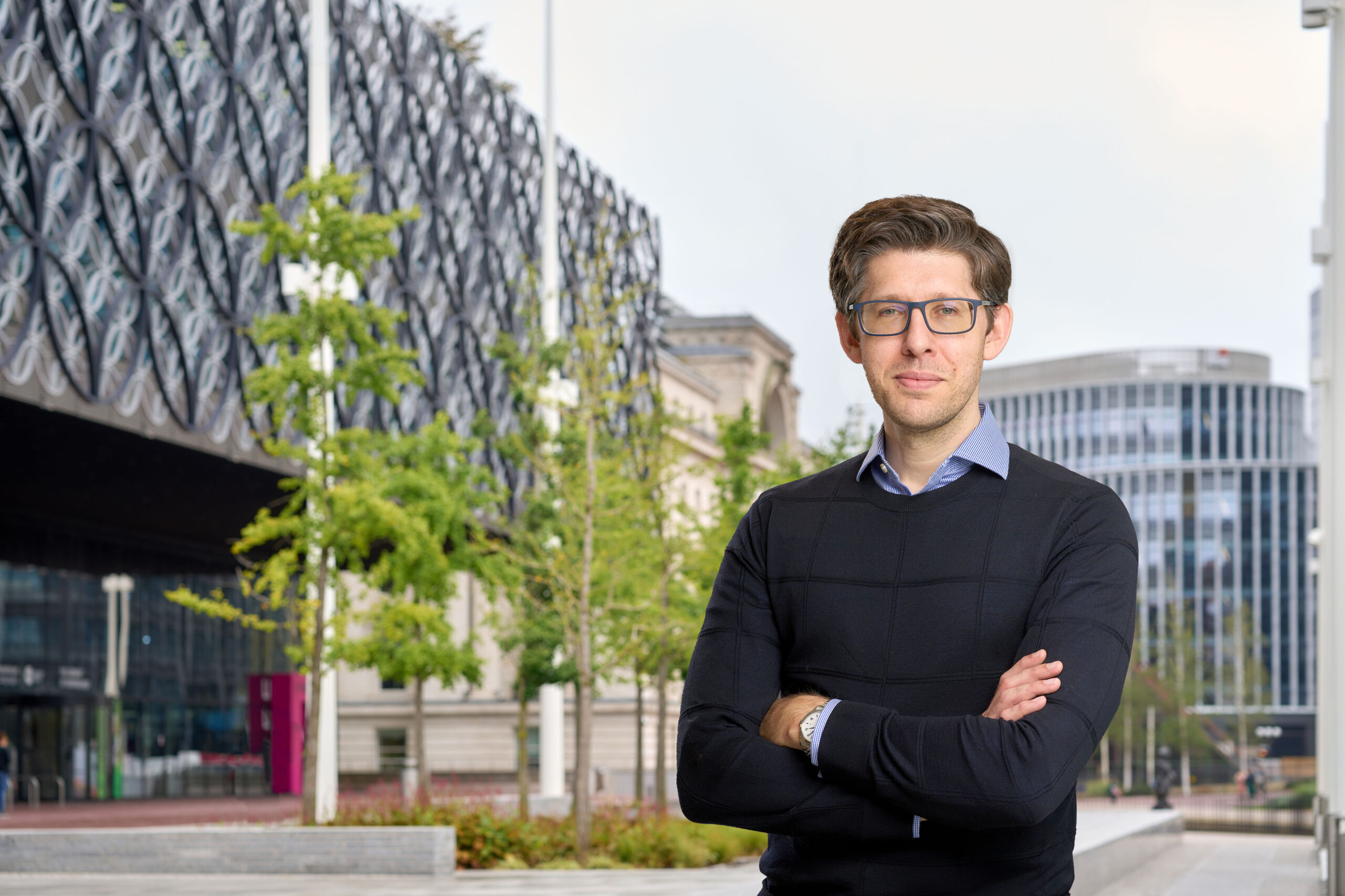 A man in a black jumper with a light blue shirt collar is standing with his arms folded in front of some trees and buildings in central Birmingham.