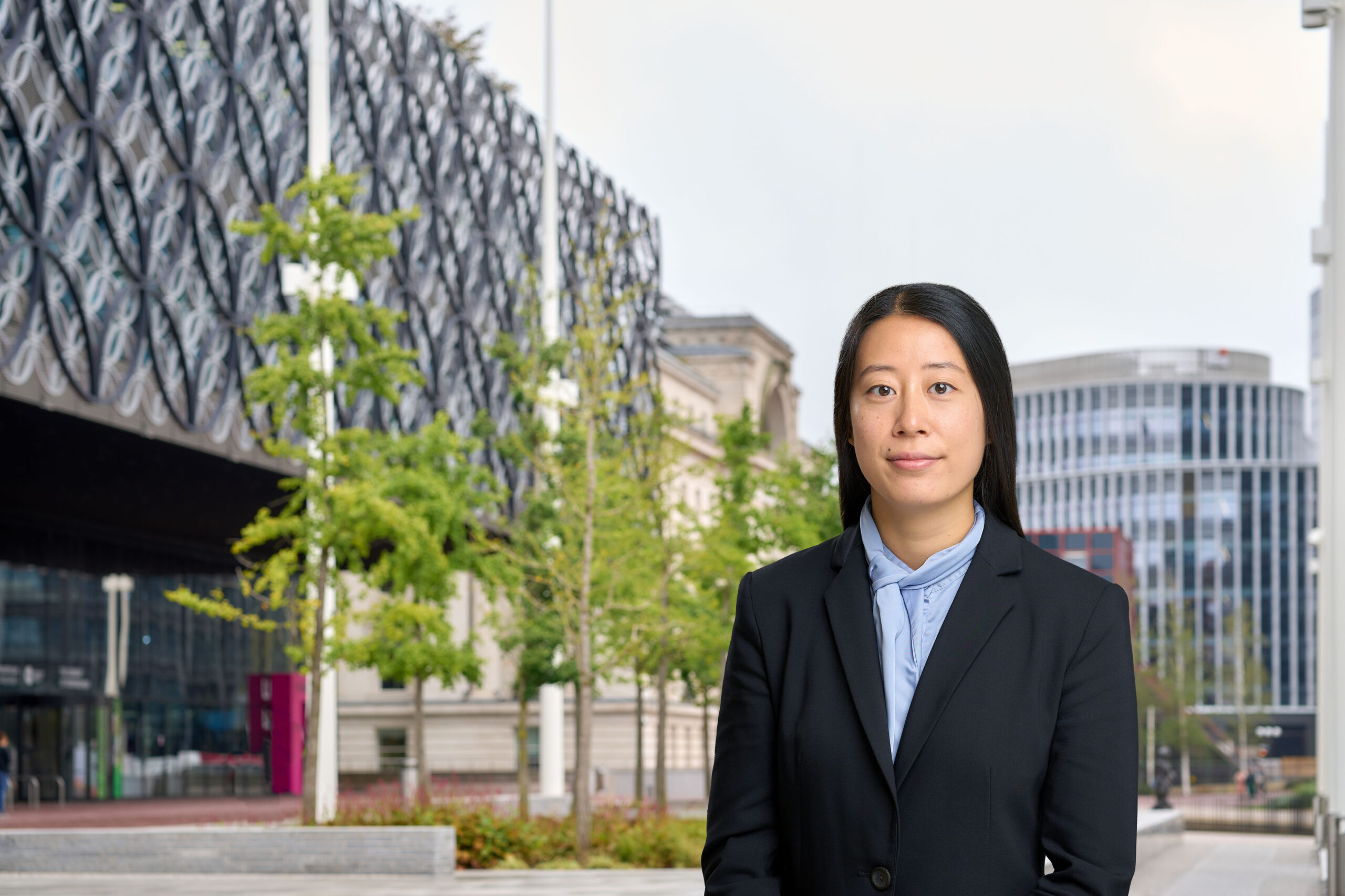 A woman in a light blue shirt and black blazer is standing in front of some trees and buildings in central Birmingham.